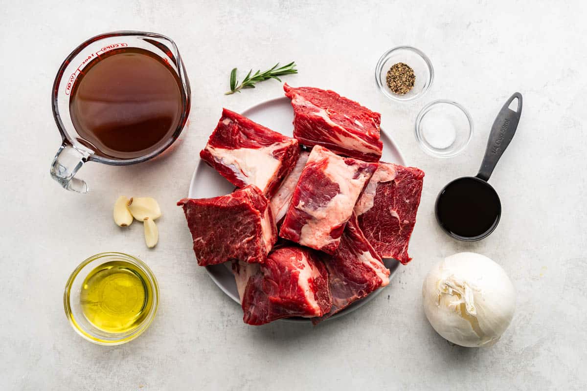 An image showing the ingredients needed to make braised beef short ribs: short ribs, beef broth, red wine, olive oil, garlic, onion, salt, pepper, and fresh rosemary.